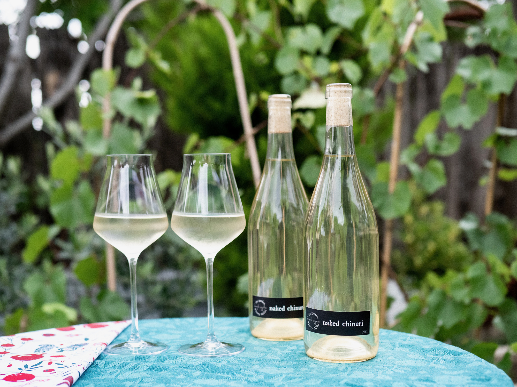 Naked Chinuri 2021 — two clear bottles and two glasses of pale wine on a garden table, vines in the background
