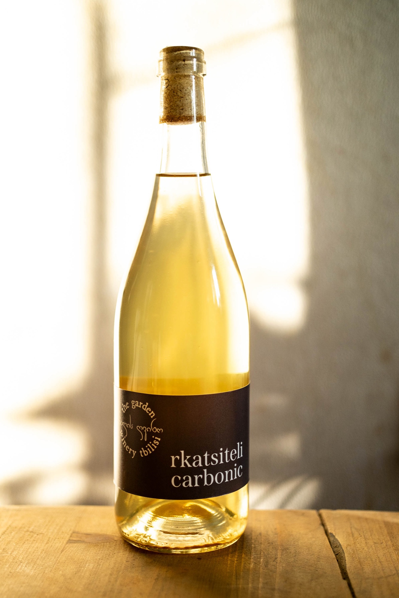 Ojaleshi Carbonic 2023 — clear bottle with teal label leaning against a tree, spring blossom alongside