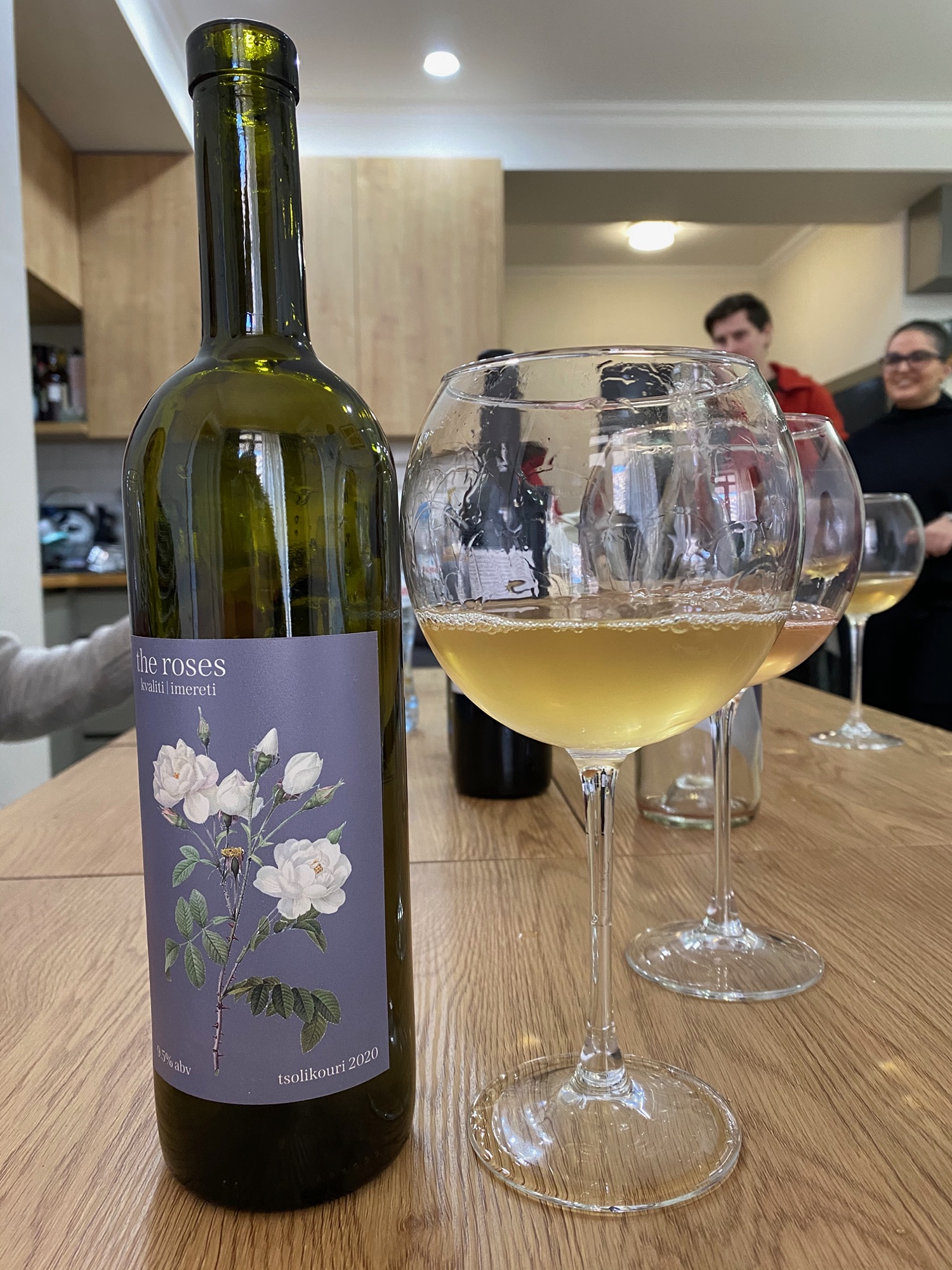 The Roses 2020 — dark bottle with botanical rose label alongside a large glass of pale wine, guests in the background